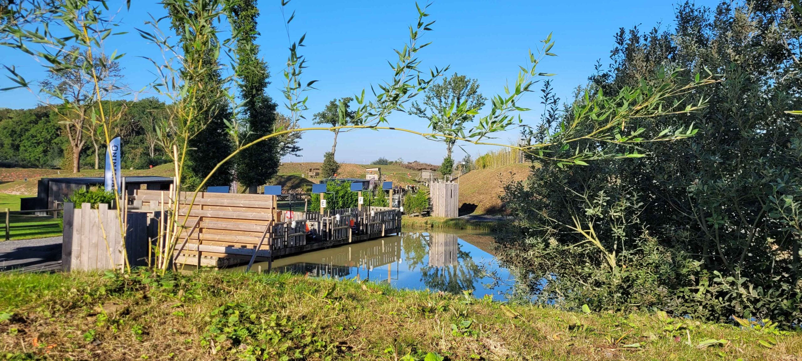 Clay shooting range with wooden shooting stands reflected in a calm pond, surrounded by green trees and blue sky, showcasing an organized outdoor shooting environment.