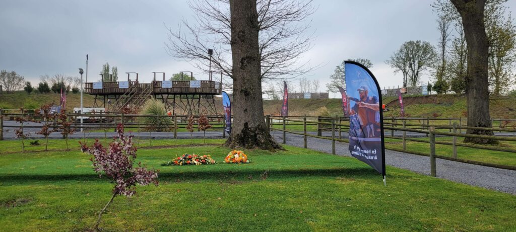 Clay shooting memorial setup with flowers under a tree, promotional Browning flag, and elevated wooden shooting platform in the background