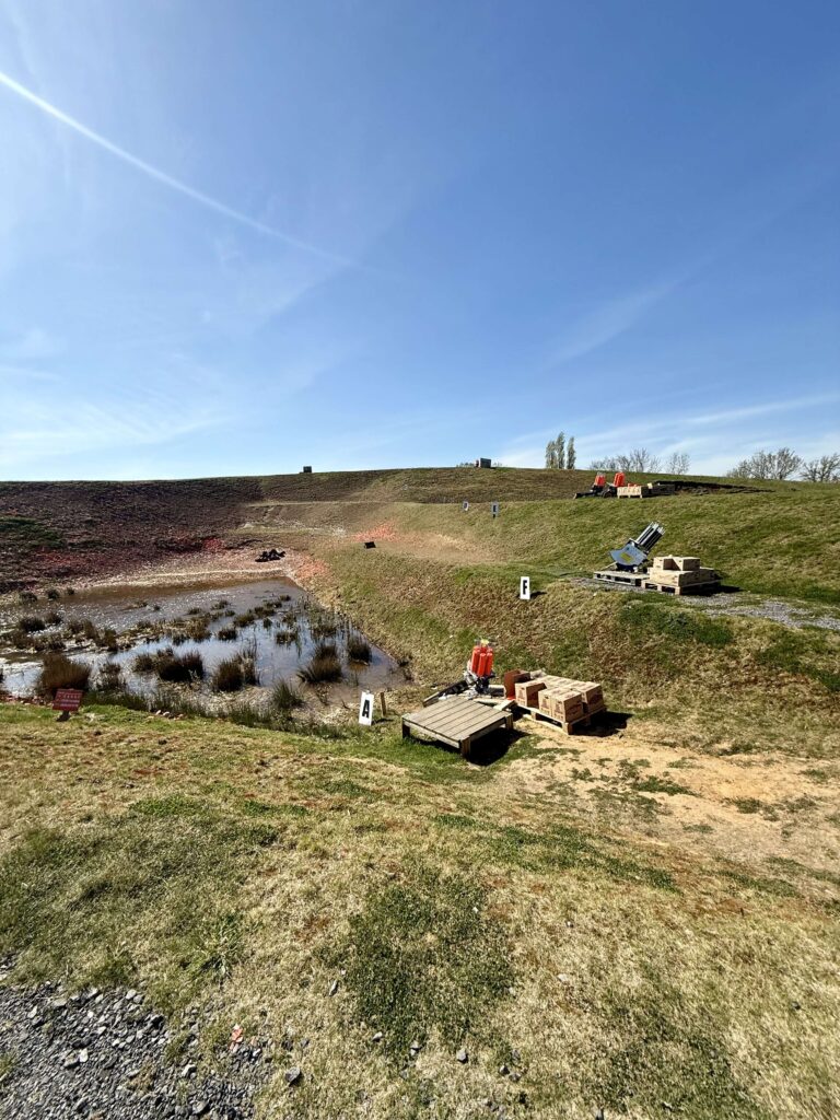 Clay shooting field with multiple trap machines set on grassy hills around a water-filled target zone, labeled shooting stations A and F.