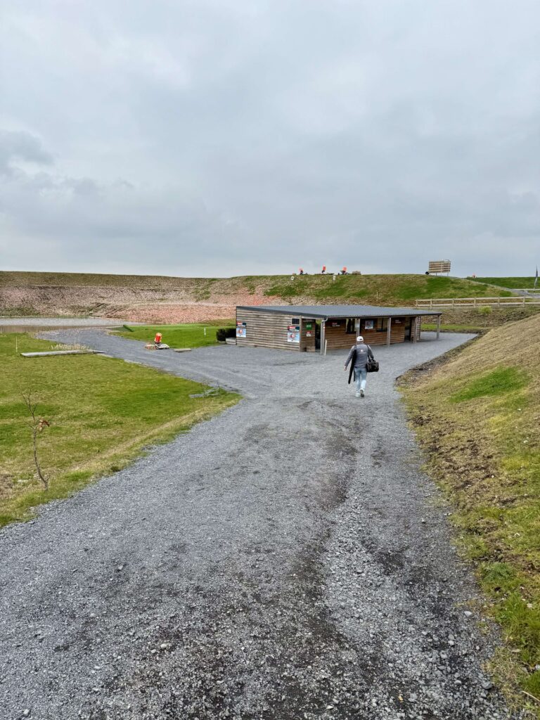 Clay shooter walking toward a wooden clubhouse on a gravel path at an outdoor shooting range with multiple clay trap machines on the hill.