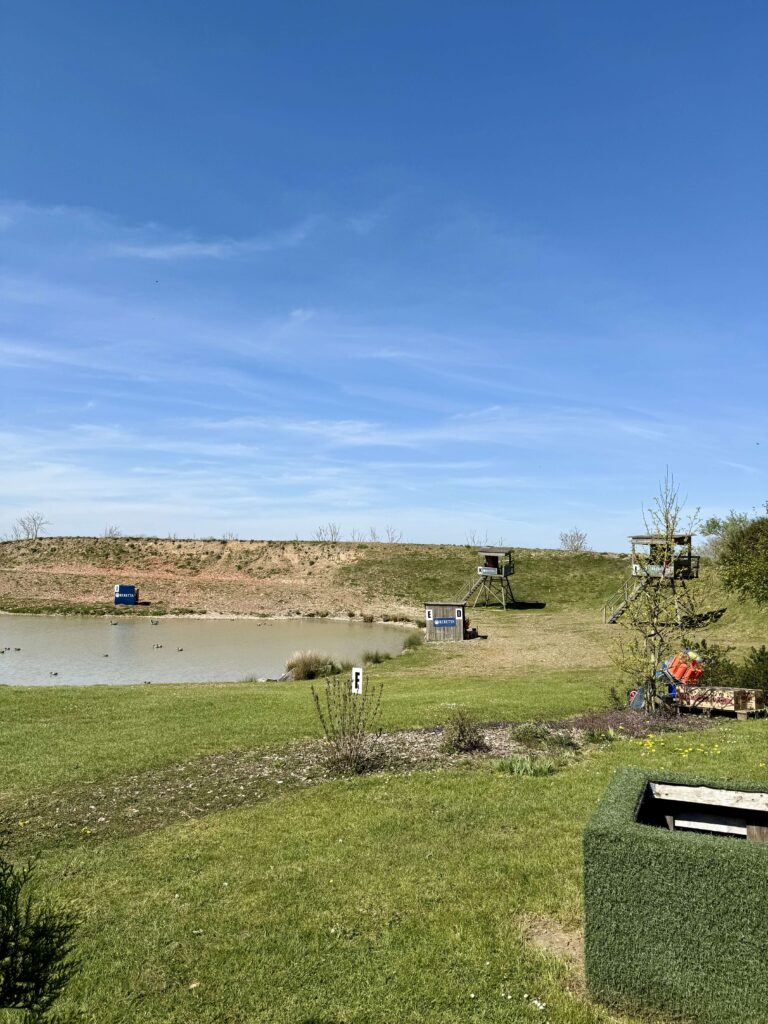 Clay shooting range with trap machines near a pond, labeled shooting station F and elevated towers in the background under a clear blue sky.