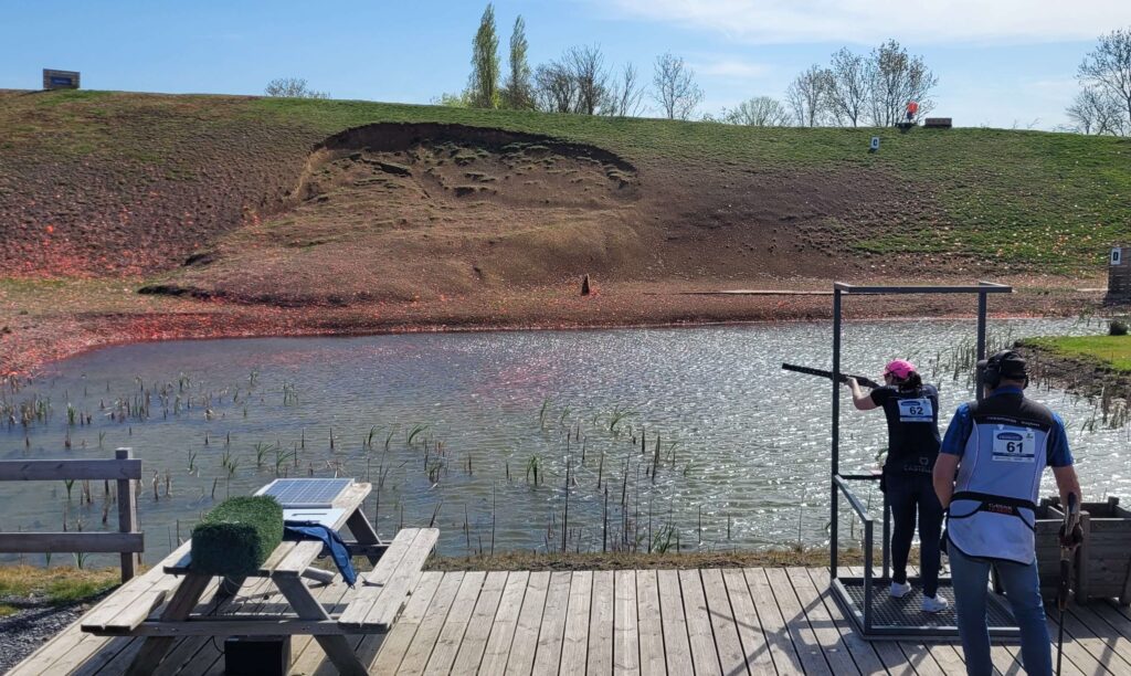Female clay shooter in a pink cap taking aim over a pond during a competition, with another competitor waiting behind her on a wooden platform
