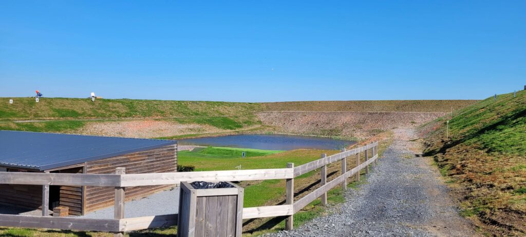 "View of a trap shooting range with multiple clay throwers on a berm, a pond in the center, and a wooden building with a gravel path in the foreground under a clear blue sky.