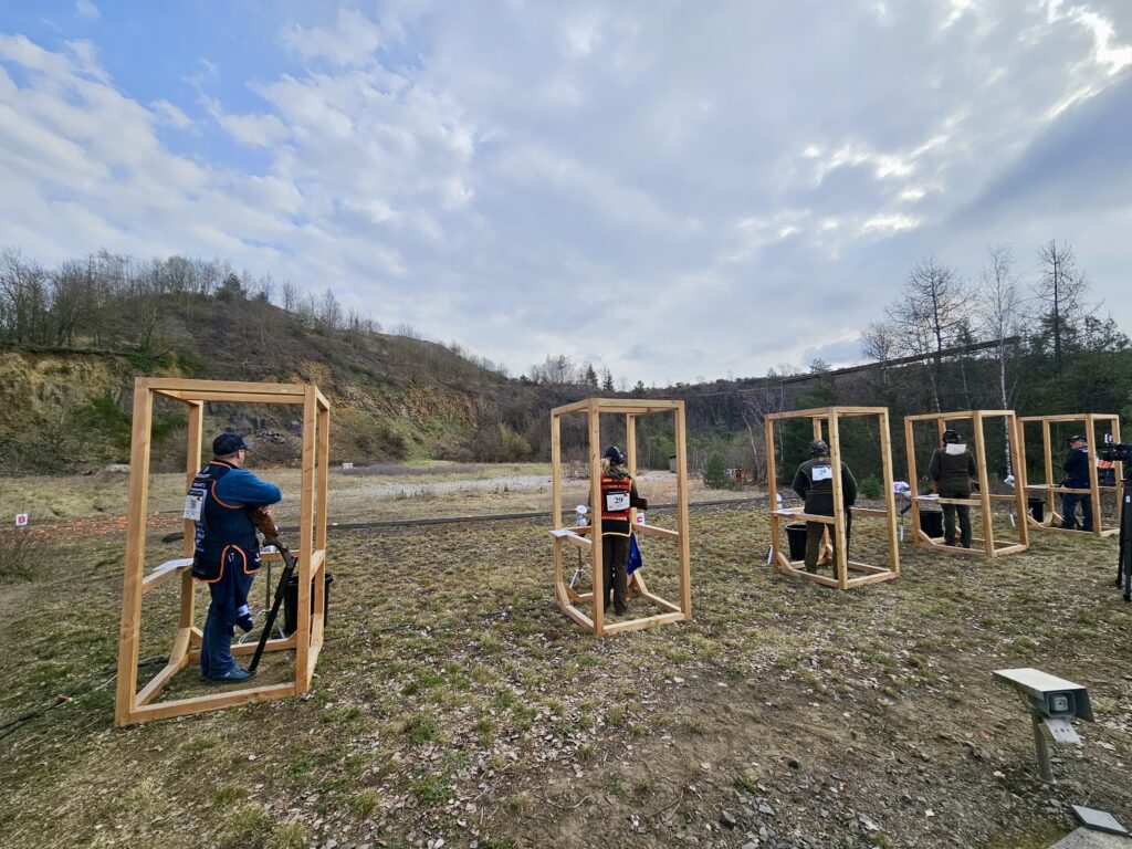 Clay shooting competitors standing in wooden shooting stations at an outdoor range during a competition, with a natural hillside backdrop and cloudy sky.