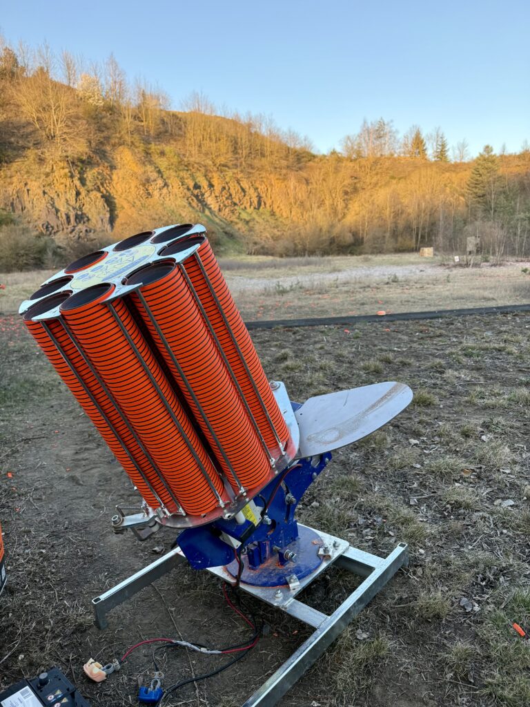 Clay target thrower machine loaded with bright orange clays, set up outdoors against a backdrop of rocky hills and leafless trees at a clay shooting range.