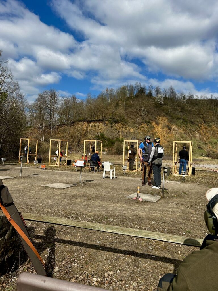 Clay shooting competitors lined up in wooden shooting stands at the Rheinland-Pfalz Cup, with coaches and spectators observing under a partly cloudy blue sky and forested hillside backdrop.